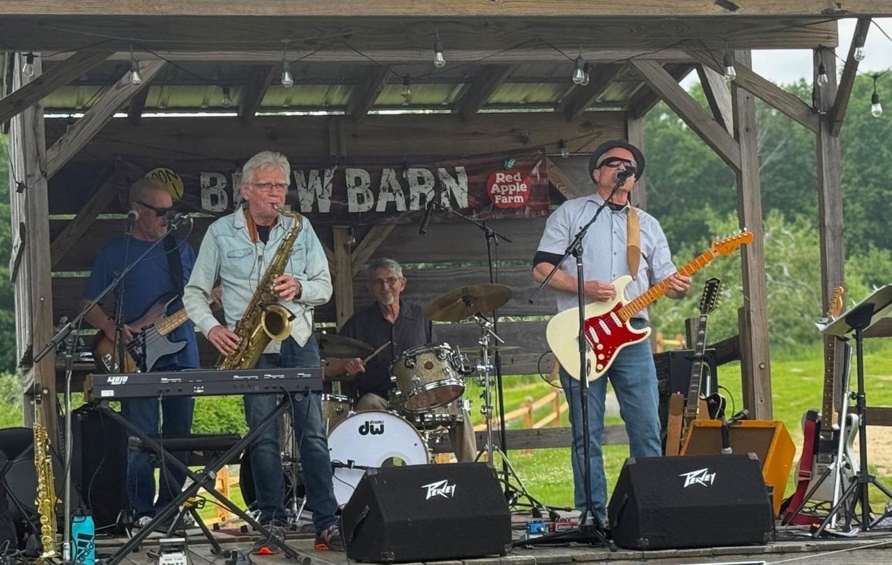The Jim Atkinson Band playing live on an outdoor wooden stage, with bass, saxophone, drums, and guitar.